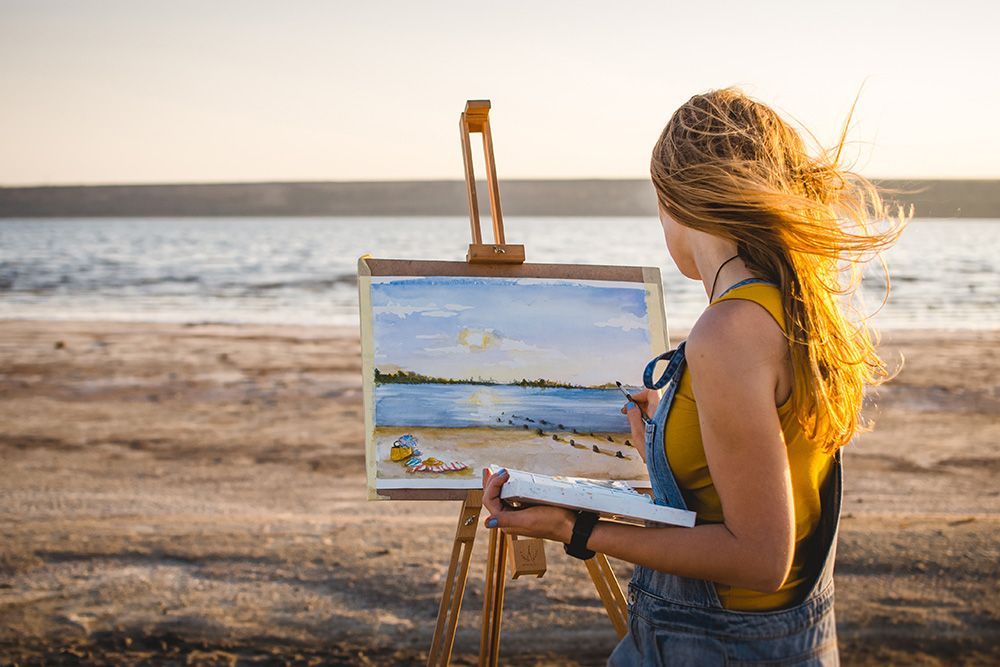 Woman paints a landscape on a beach at sunset, holding a palette and brush, with the sea behind.