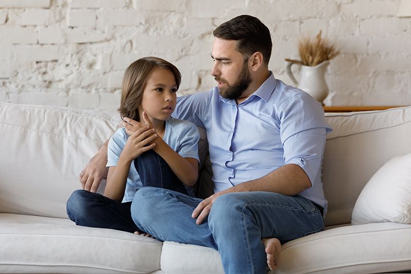 Man and child sit close on a white couch. Man has arm around child's shoulder, talking. Soft, indoor setting.