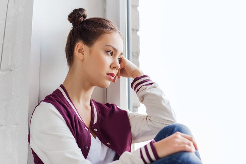 Woman with bun, sitting by a window, looking pensive. Wearing a white and maroon jacket and jeans.