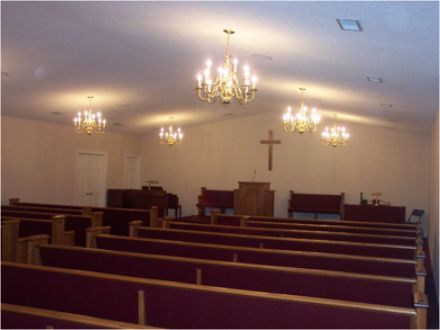 Interior of a church sanctuary with rows of pews, a cross, and chandeliers.