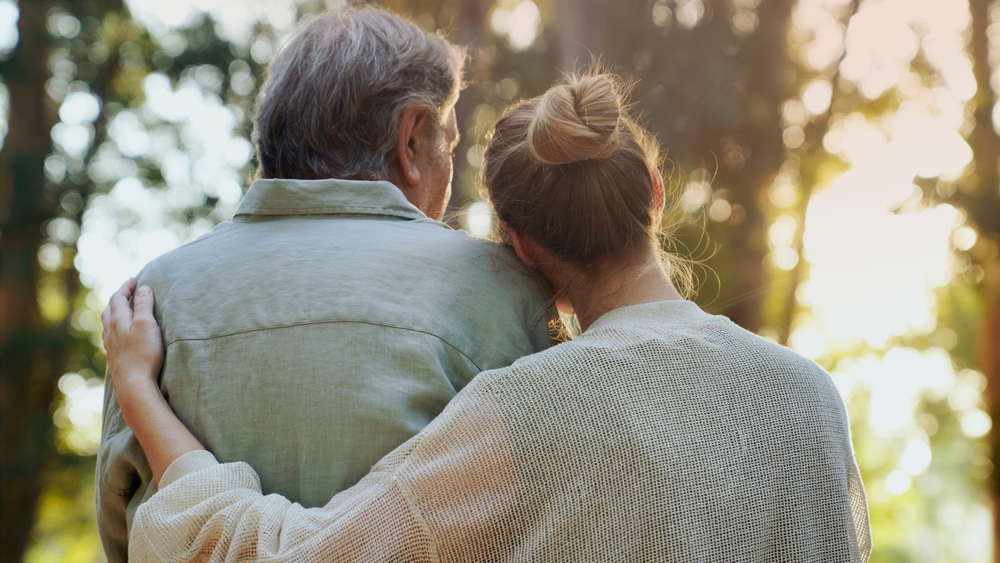 Woman comforting person outdoors, arm around their shoulder. Sunlight filters through trees.