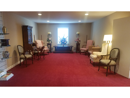 Funeral home viewing room with red carpet, neutral walls, chairs, and a floral arrangement.