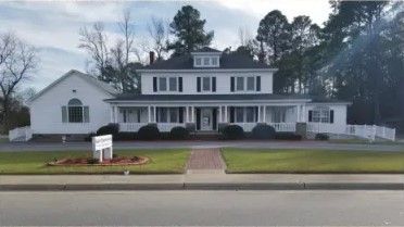 White two-story house with wrap-around porch, brick walkway, and a small sign in front on a green lawn.