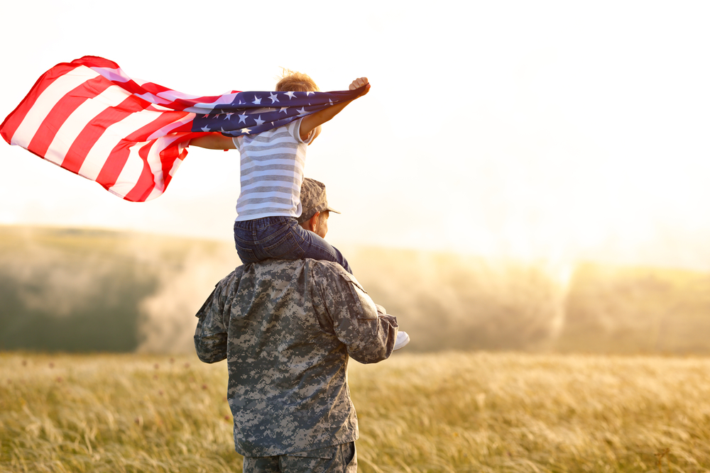 Soldier carrying child on shoulders, waving American flag in field at sunset.