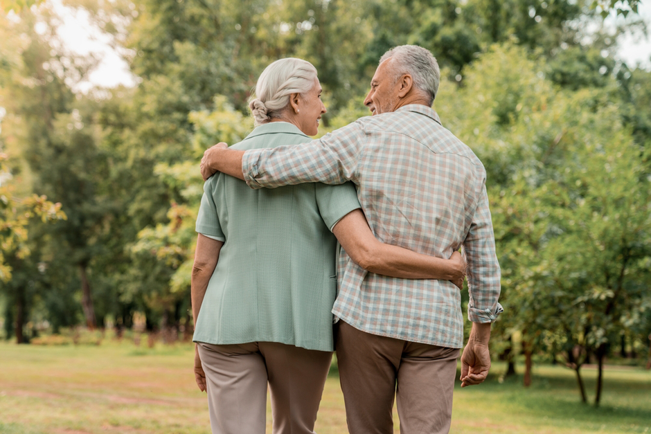 Elderly couple with arms around each other, walking in a park with trees, backs to the camera.