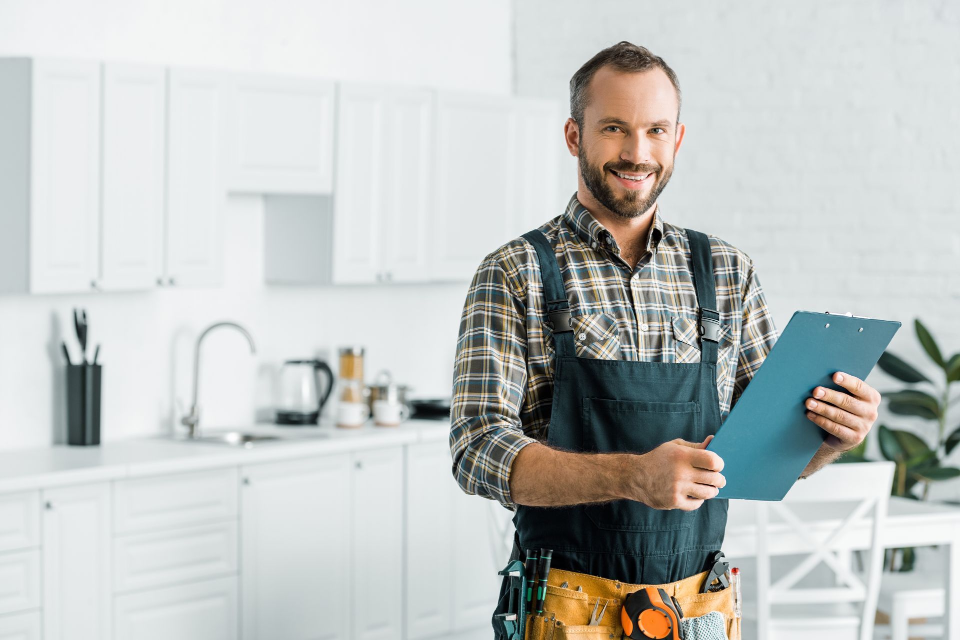 Home repair worker with tool belt holding clipboard in modern kitchen interior.