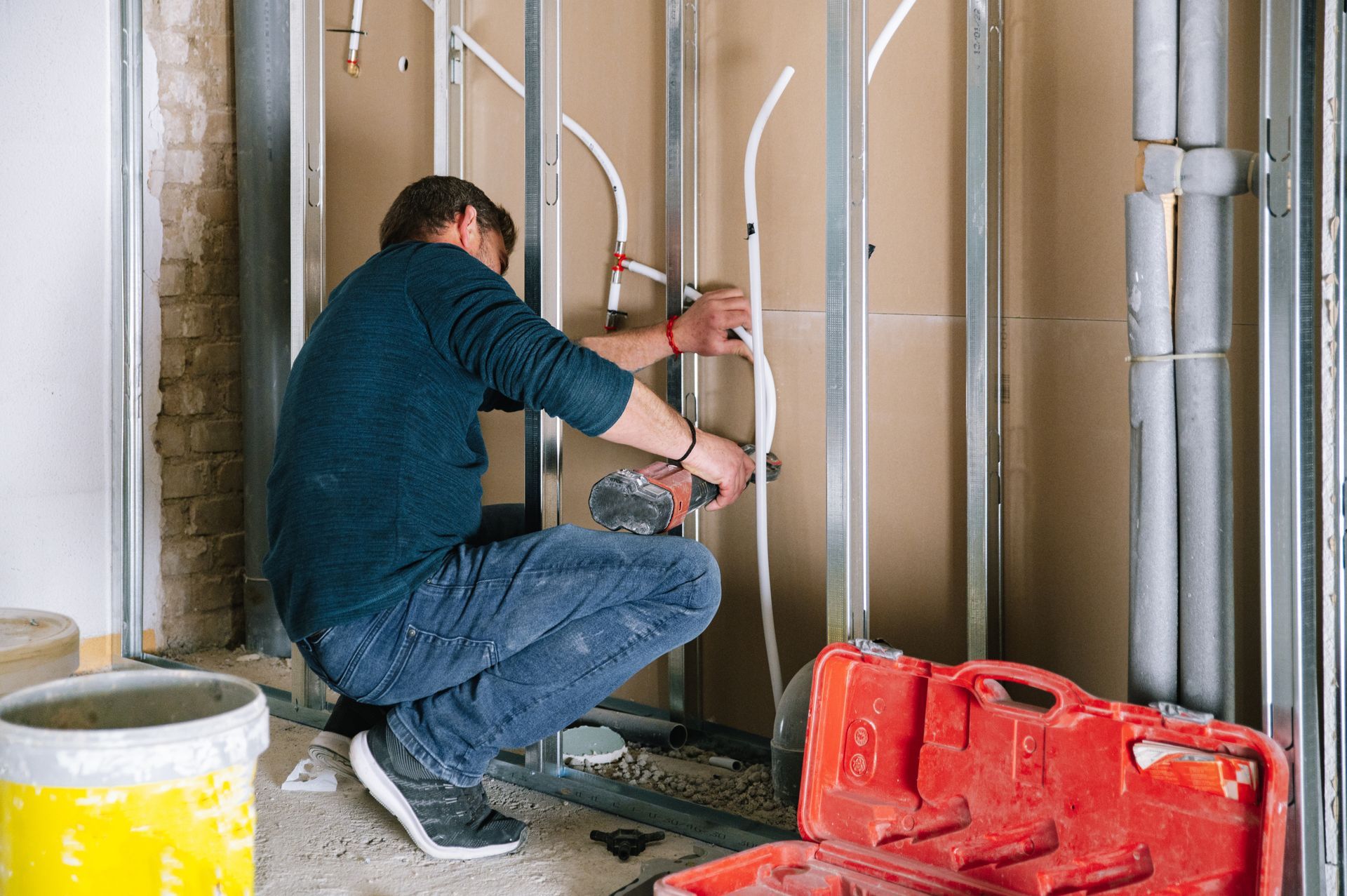 Worker installing plumbing pipes inside a building under renovation, with tools and buckets nearby