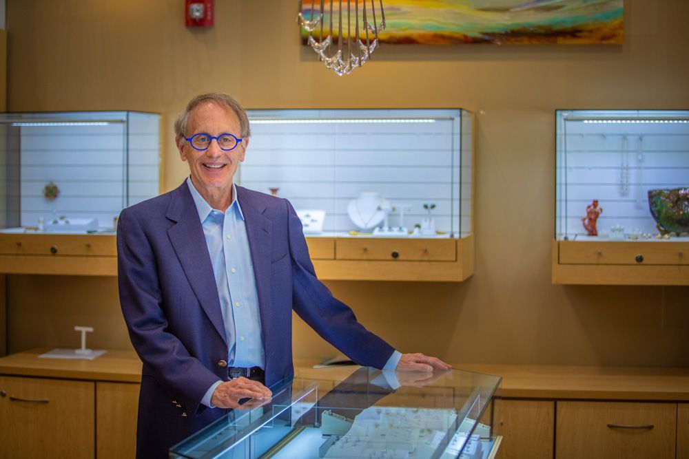 Man standing behind a glass display case in a warmly lit gallery or showroom, smiling at the camera