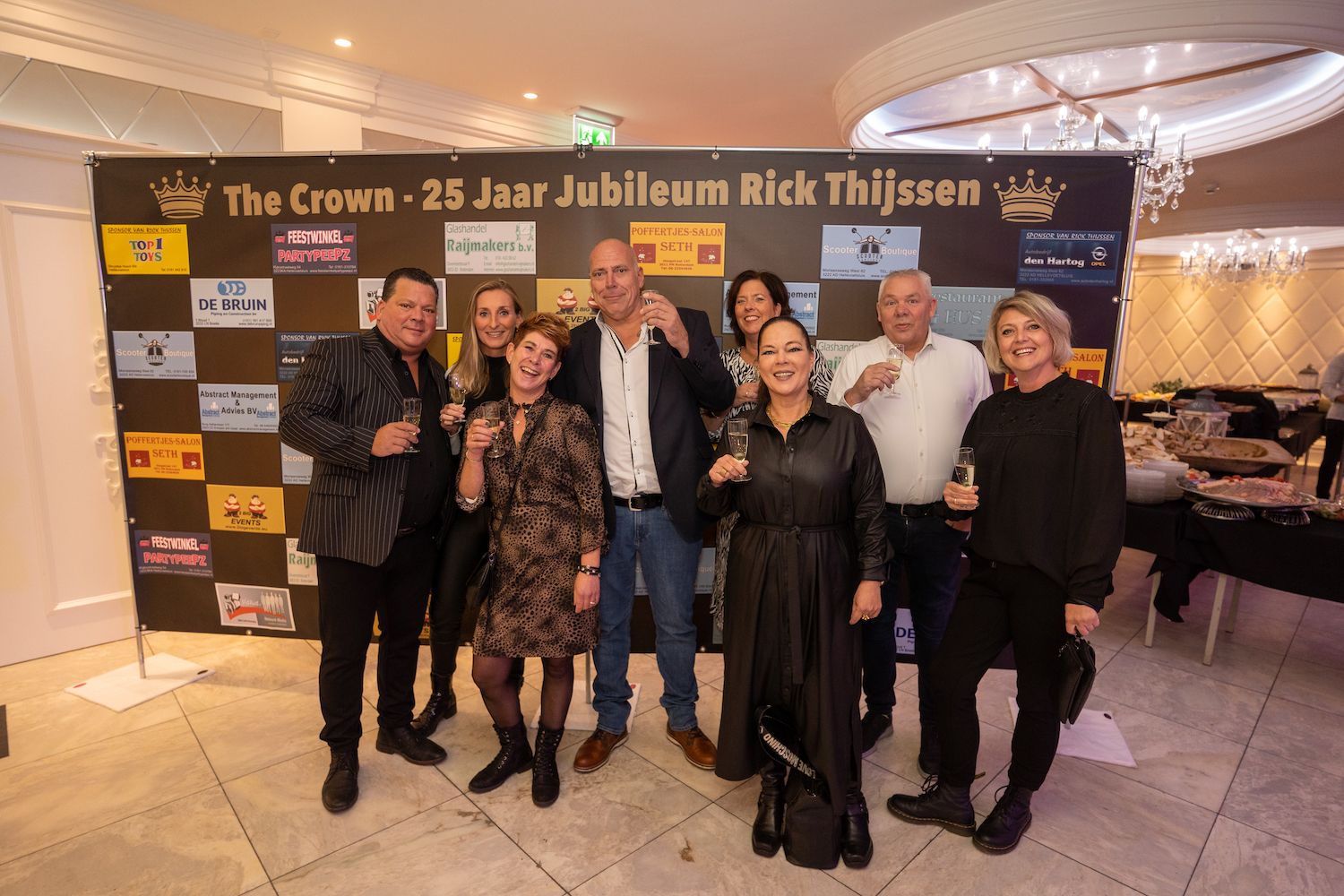 Zes mensen poseren tijdens een jubileumevenement met een zwart-gouden banner op de achtergrond.