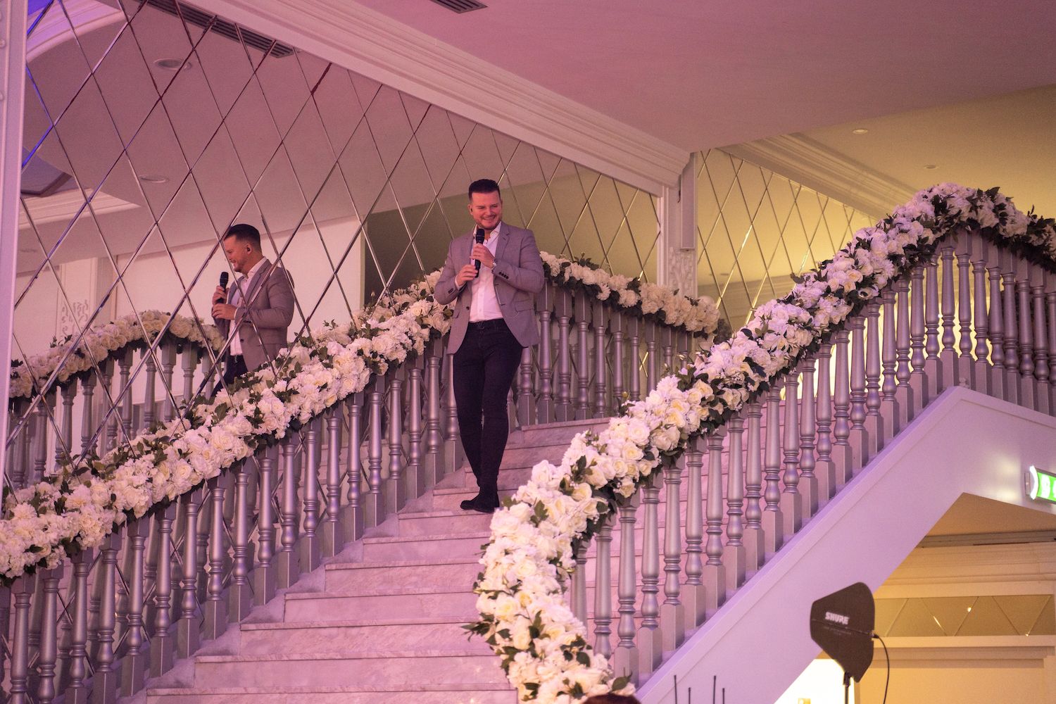 Twee mannen dalen een met bloemen versierde trap af in een rijkversierde evenementenlocatie.