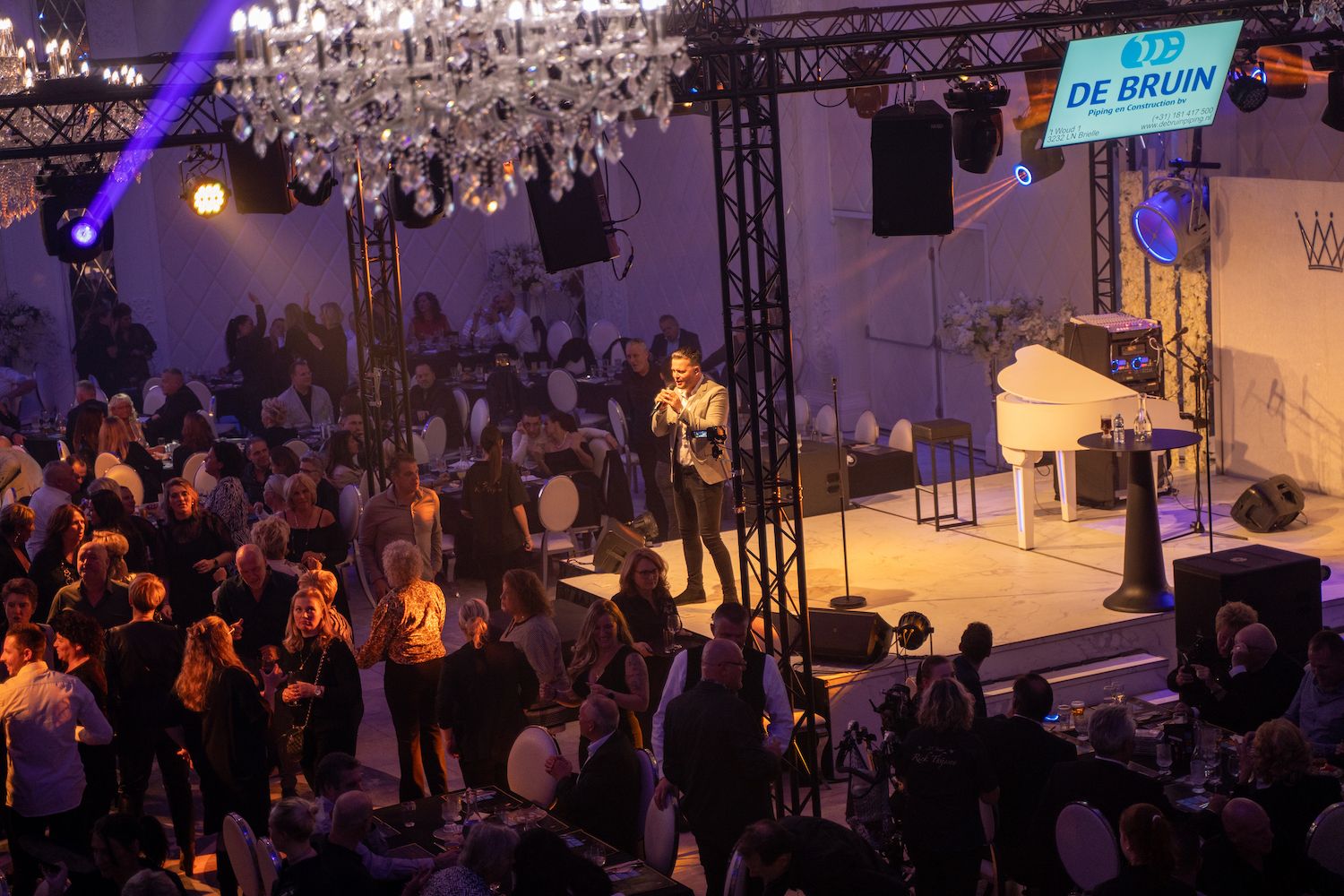 Een drukbezocht indoorconcert met artiesten op het podium onder paarse lichten en een witte piano.