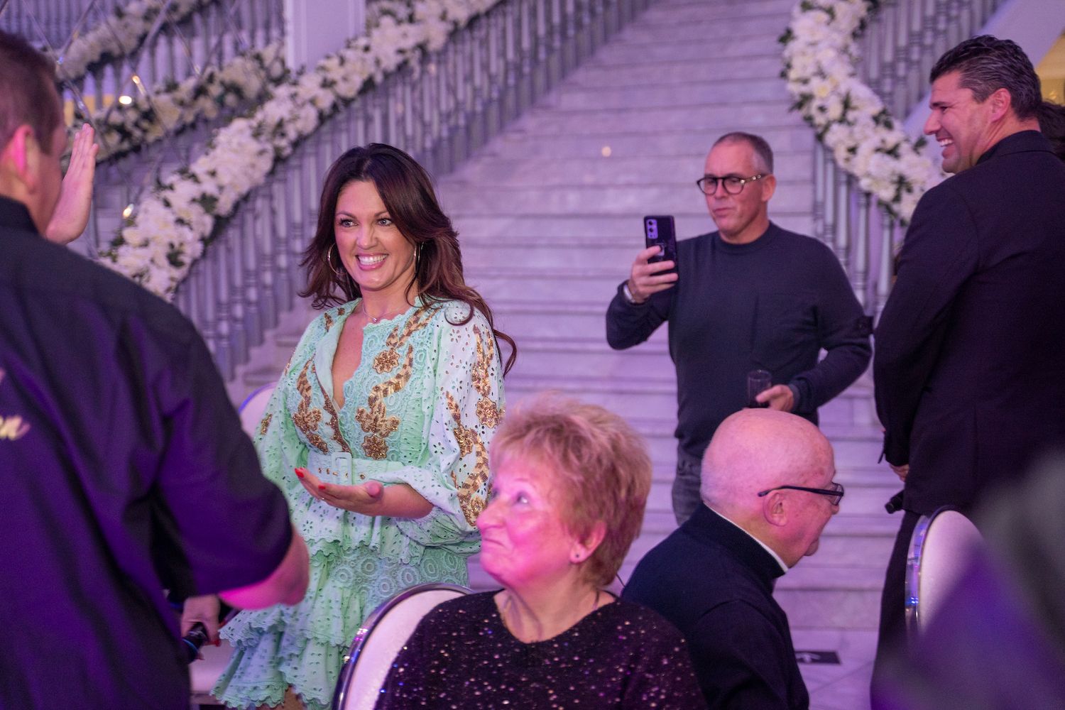 Gasten die zich mengen op een formeel evenement, met een lachende vrouw in een mintgroene jurk bij een trap en bloemversieringen.