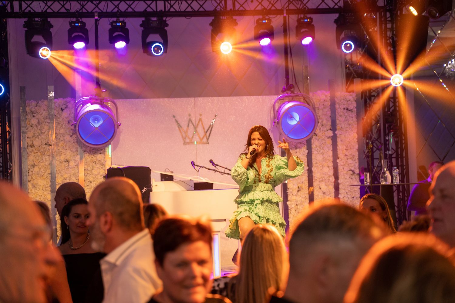 Zangeres treedt op een verlicht podium op tijdens een evenement binnenshuis, met publiek op de voorgrond.