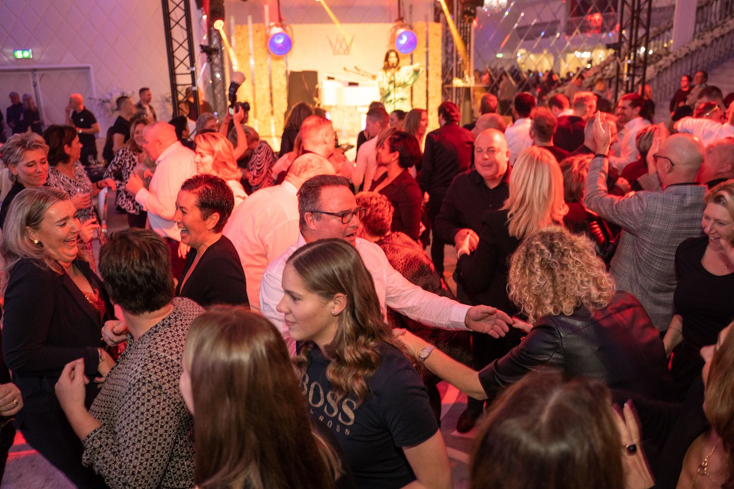 Een drukbezocht binnenfeest met mensen die dansen onder warme rode en paarse lichten.