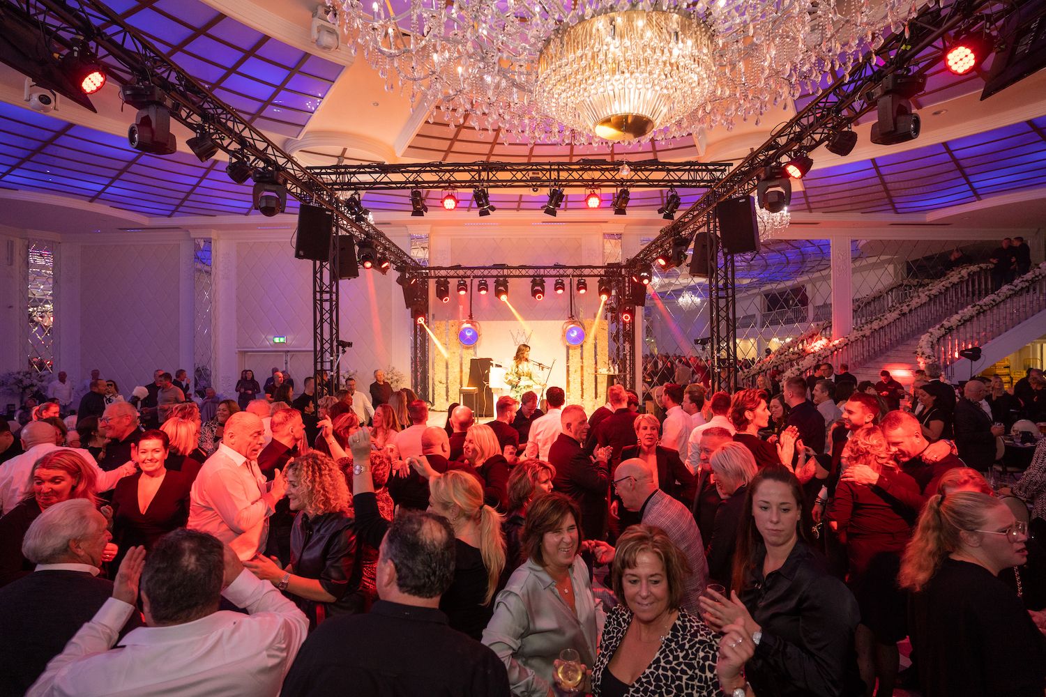 Een drukbezochte indoor gala met mensen die dansen onder kroonluchters en roze-paarse podiumverlichting.