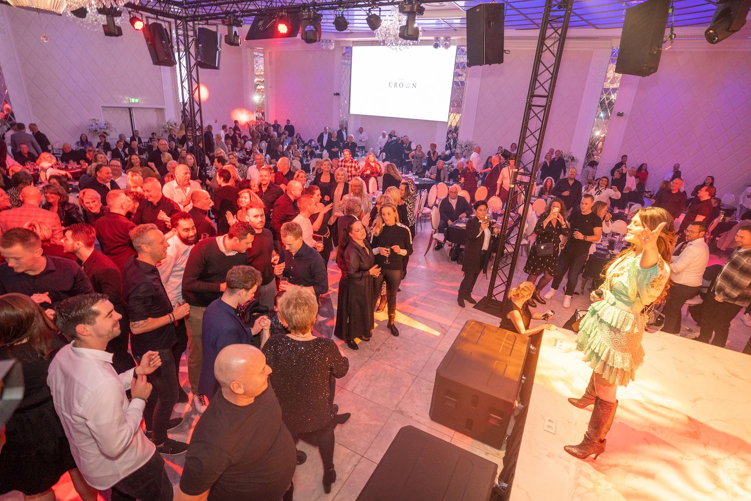 Een drukbezocht evenement binnenshuis, waar mensen naar een artiest op een podium onder roze verlichting kijken.