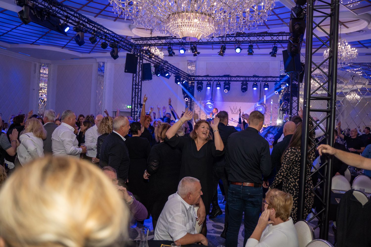 Een drukbezocht binnenfeest met mensen die dansen onder blauwe verlichting en kroonluchters.