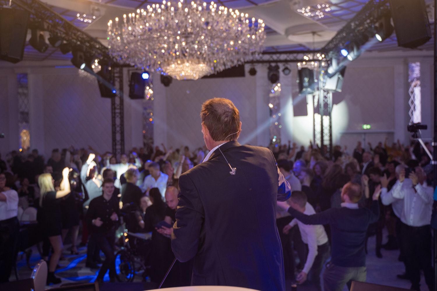 De spreker staat tegenover een volle balzaal onder kroonluchters, terwijl de gasten hun handen opsteken en dansen.