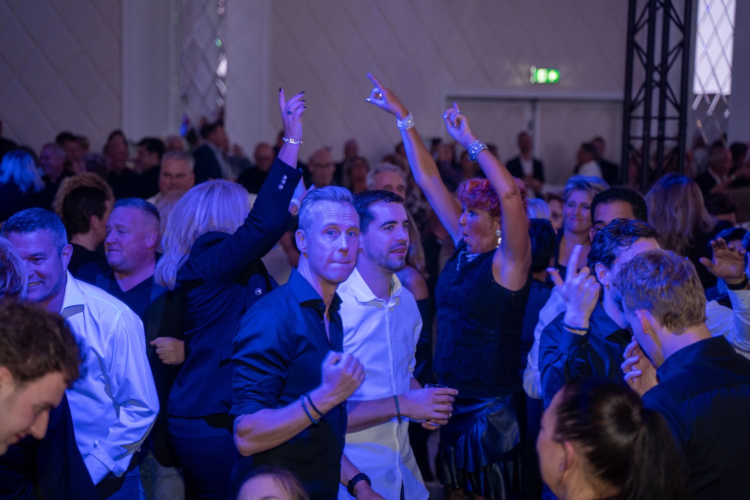 Een menigte danst op een feest onder blauwe verlichting, terwijl mensen hun drankjes heffen en op de muziek bewegen.