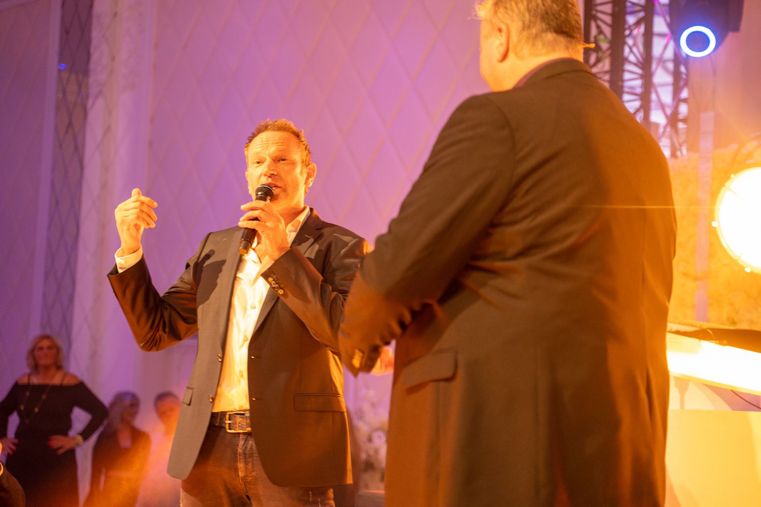 Twee mannen spreken op een podium met microfoons, een van hen kijkt naar het publiek onder warm oranje licht.