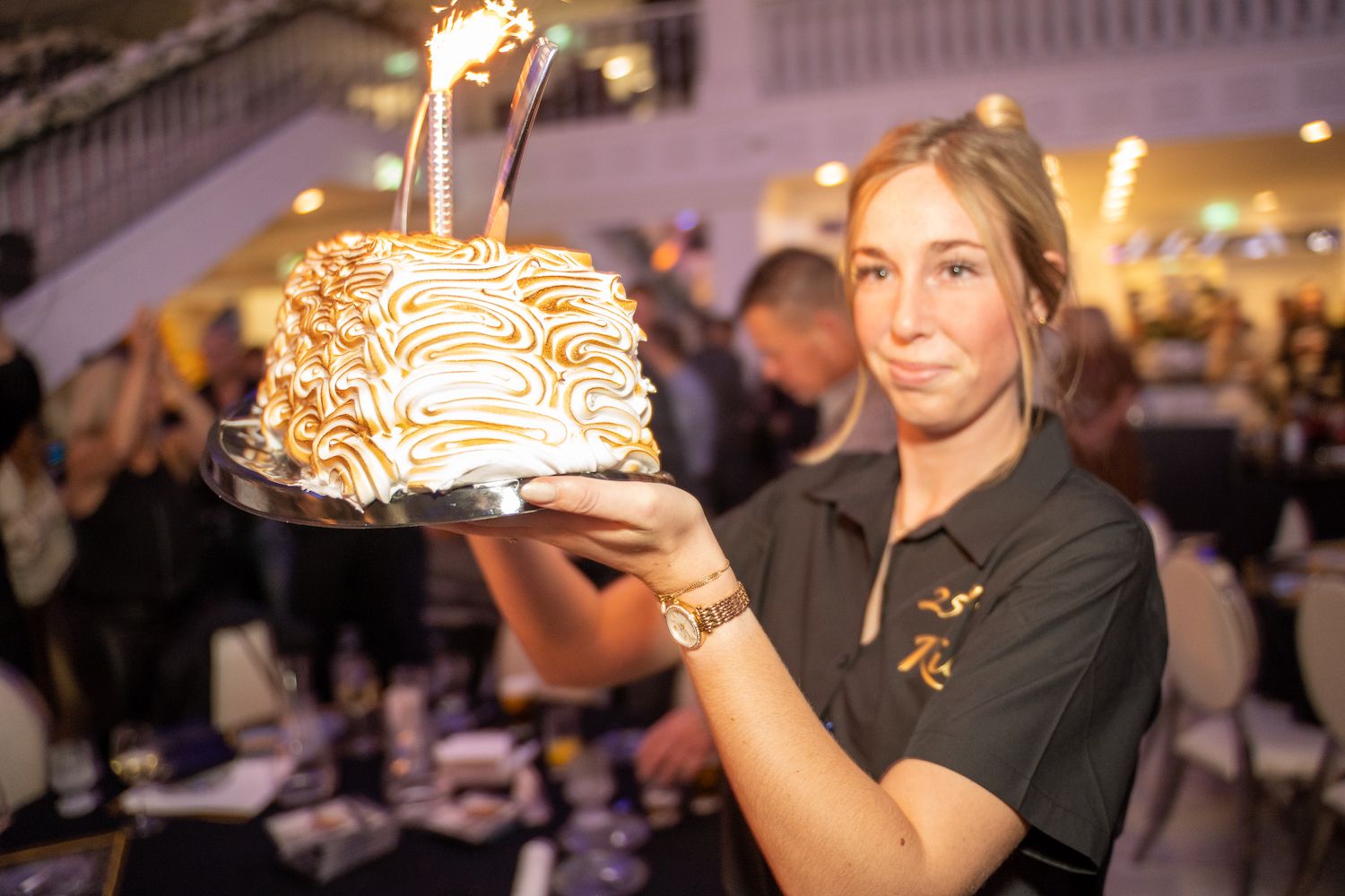 Een vrouw houdt een vlammend dessert vast tijdens een evenement binnenshuis, met een menigte en tafels op de achtergrond.