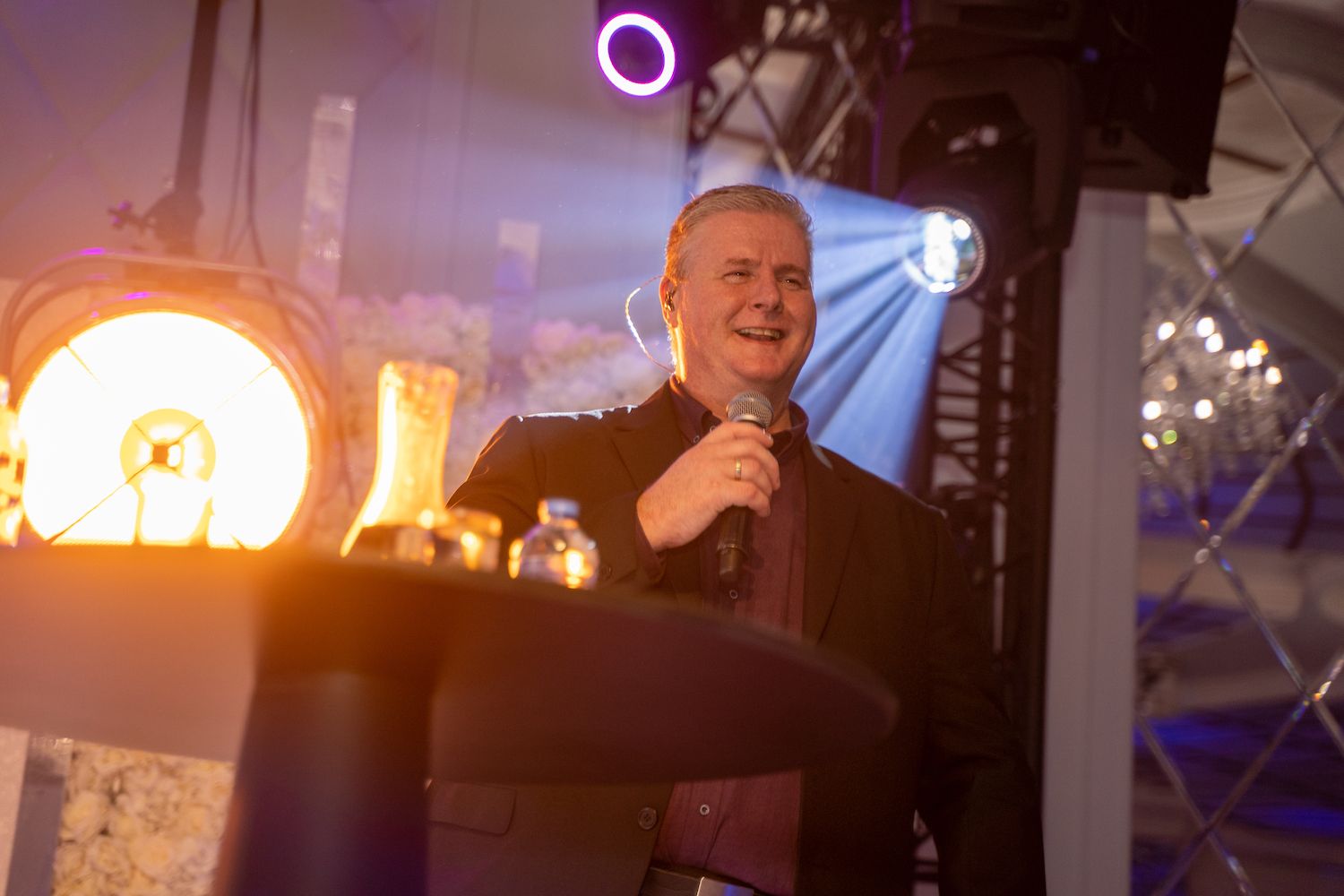 Een man met een microfoon op een podium met paarse lichten en een fel ronde schijnwerper achter hem.