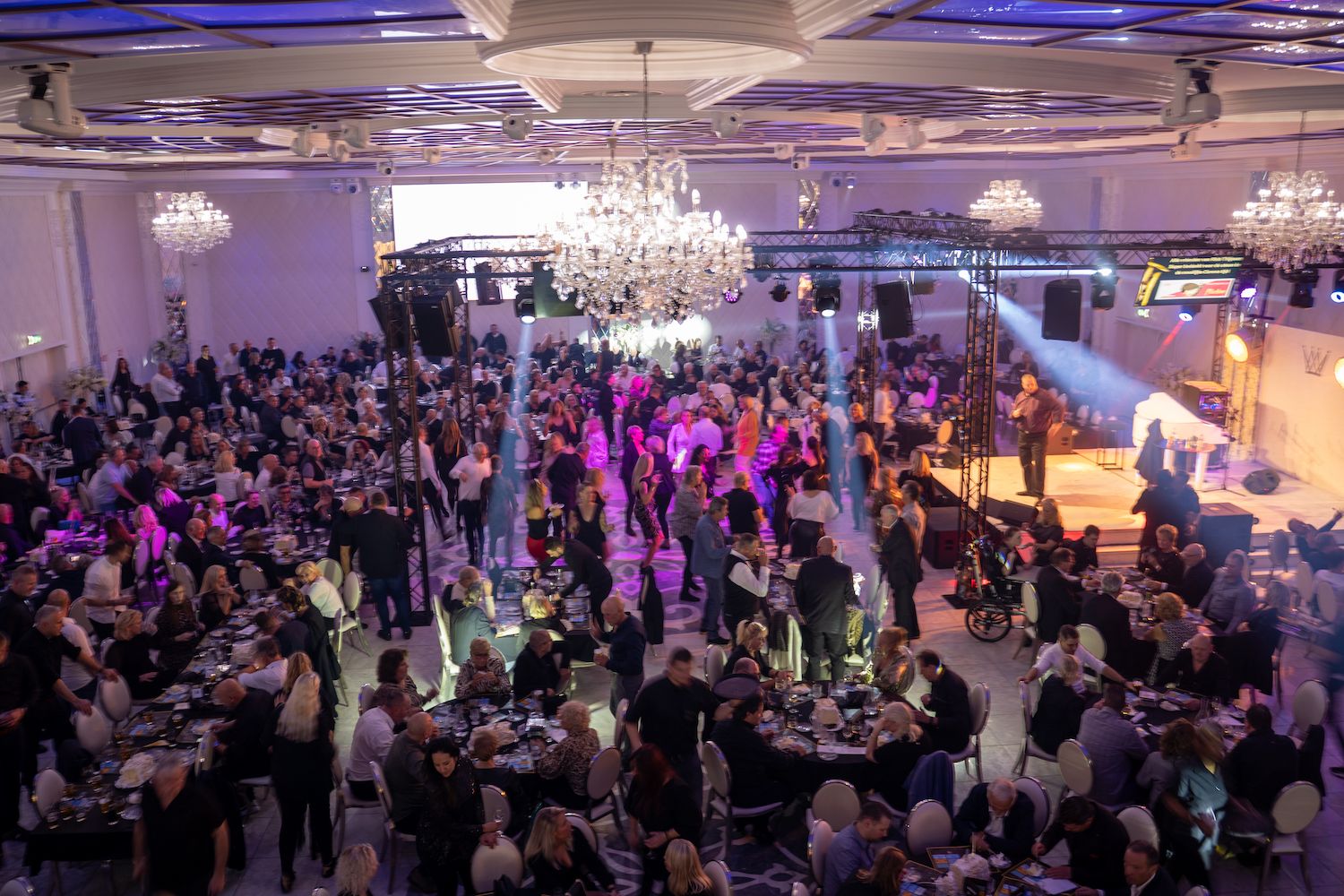 Een overvolle feestzaal met kroonluchters, paarse verlichting en gasten die aan ronde tafels zitten en dansen.