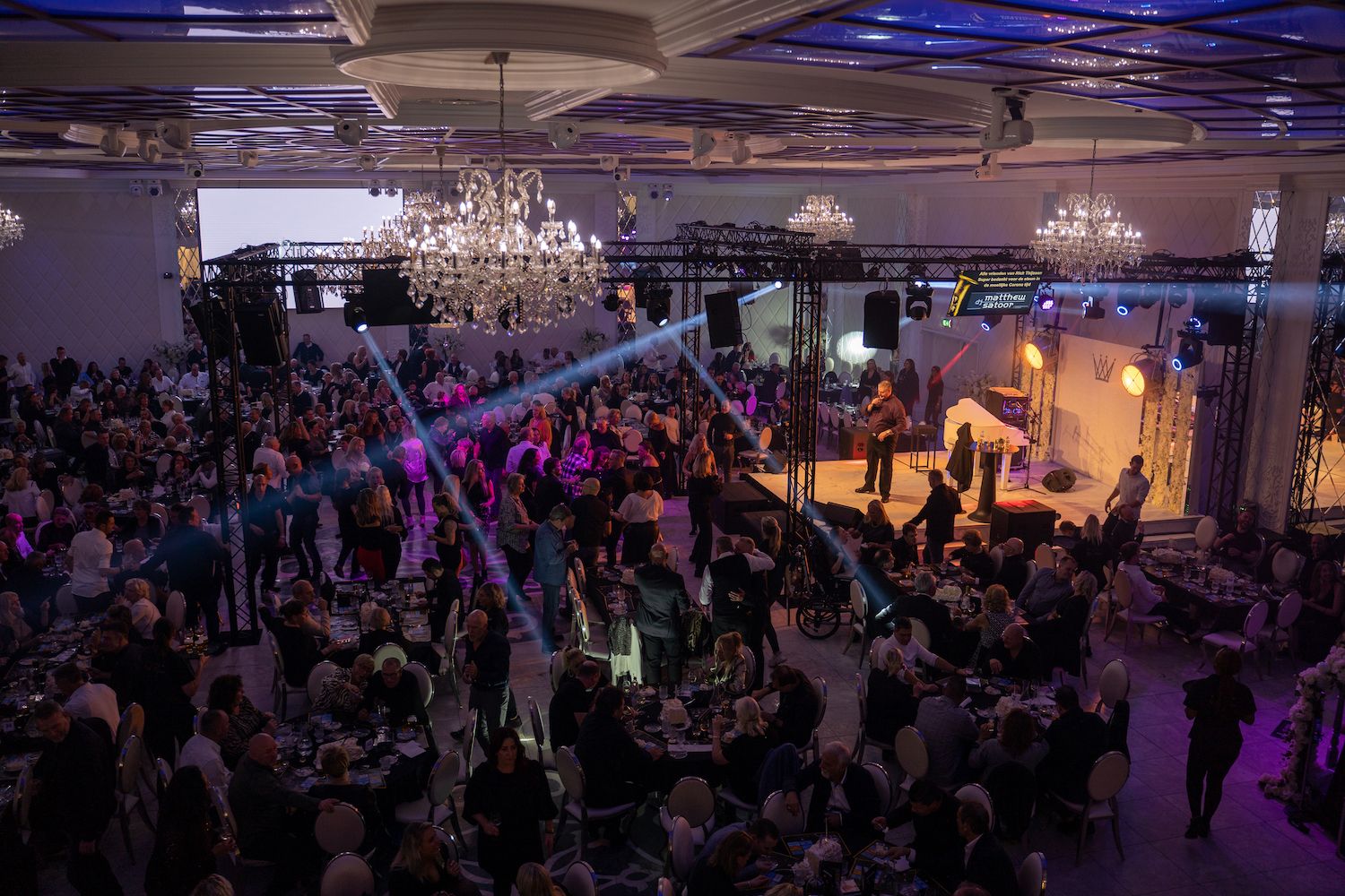 Een volle balzaal met een live band die optreedt op een klein podium onder kroonluchters en paarse verlichting.