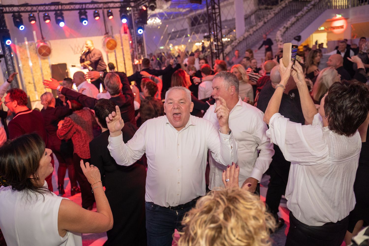 Mensen dansen op een levendig binnenfeest met podiumverlichting en opgeheven handen.