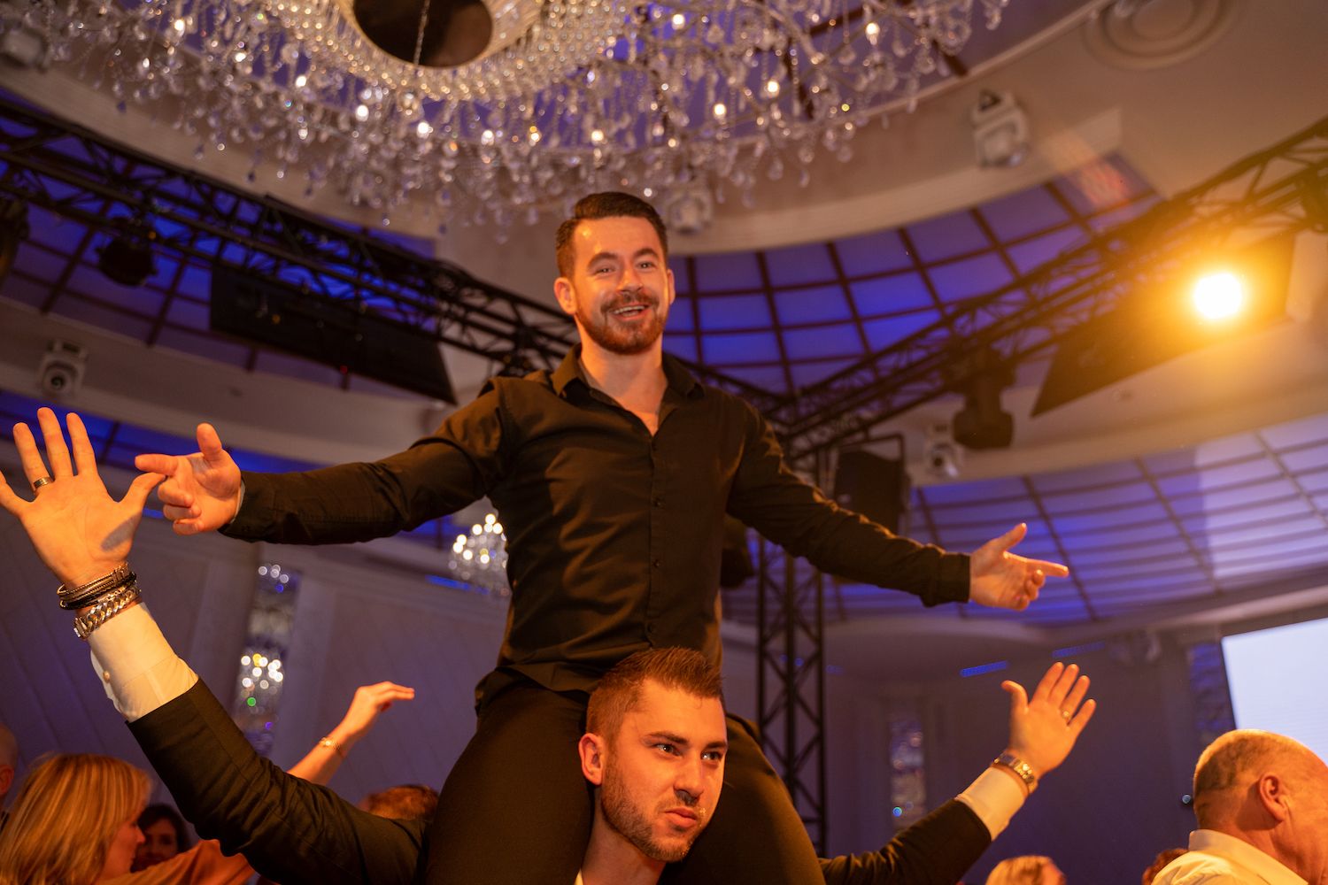 Een man glimlacht op de schouder van een ander in een drukke, warm verlichte feestzaal, met uitgestrekte armen.