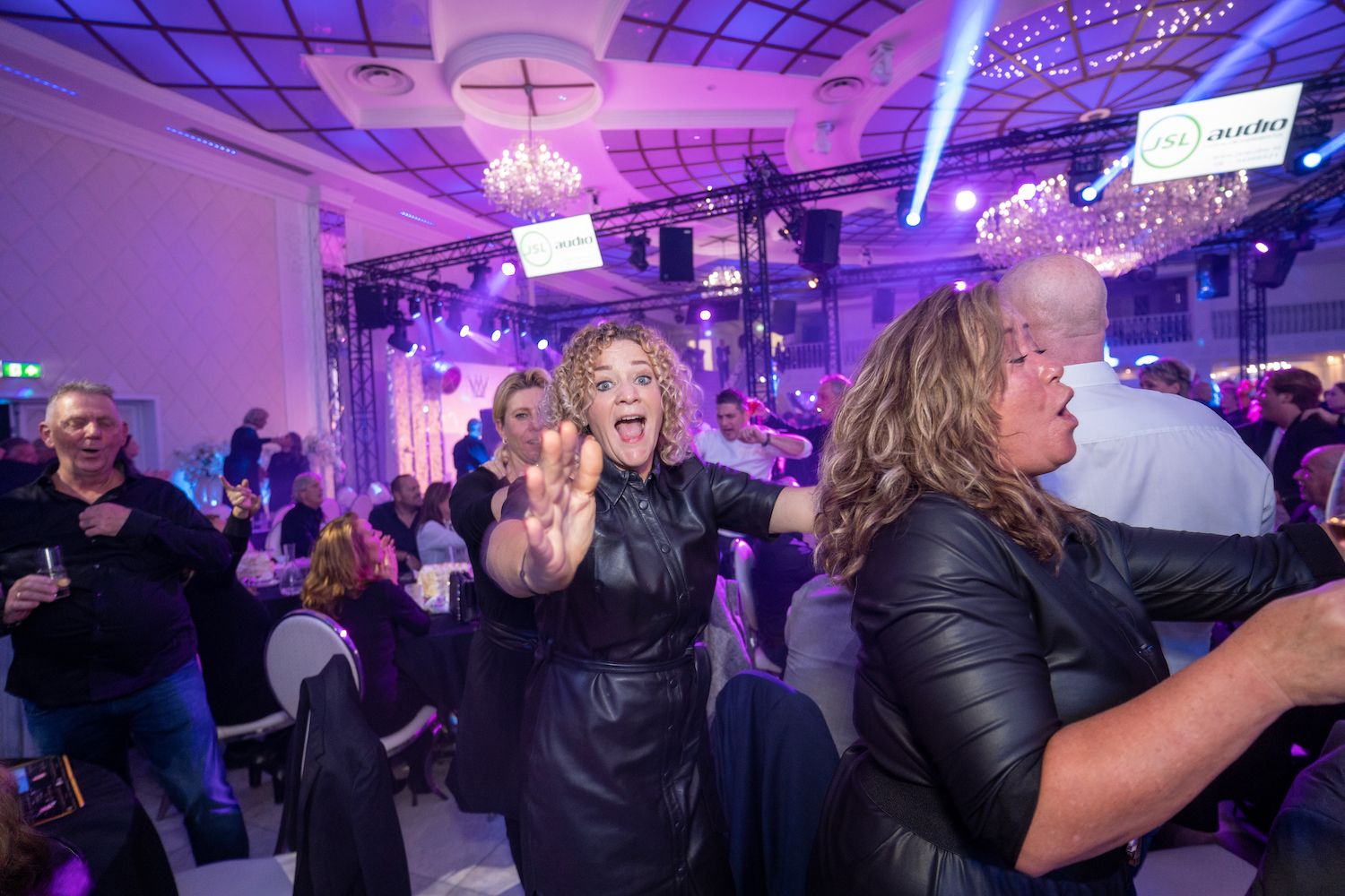 Mensen dansen en juichen onder paarse lichten op een levendig binnenfeest.