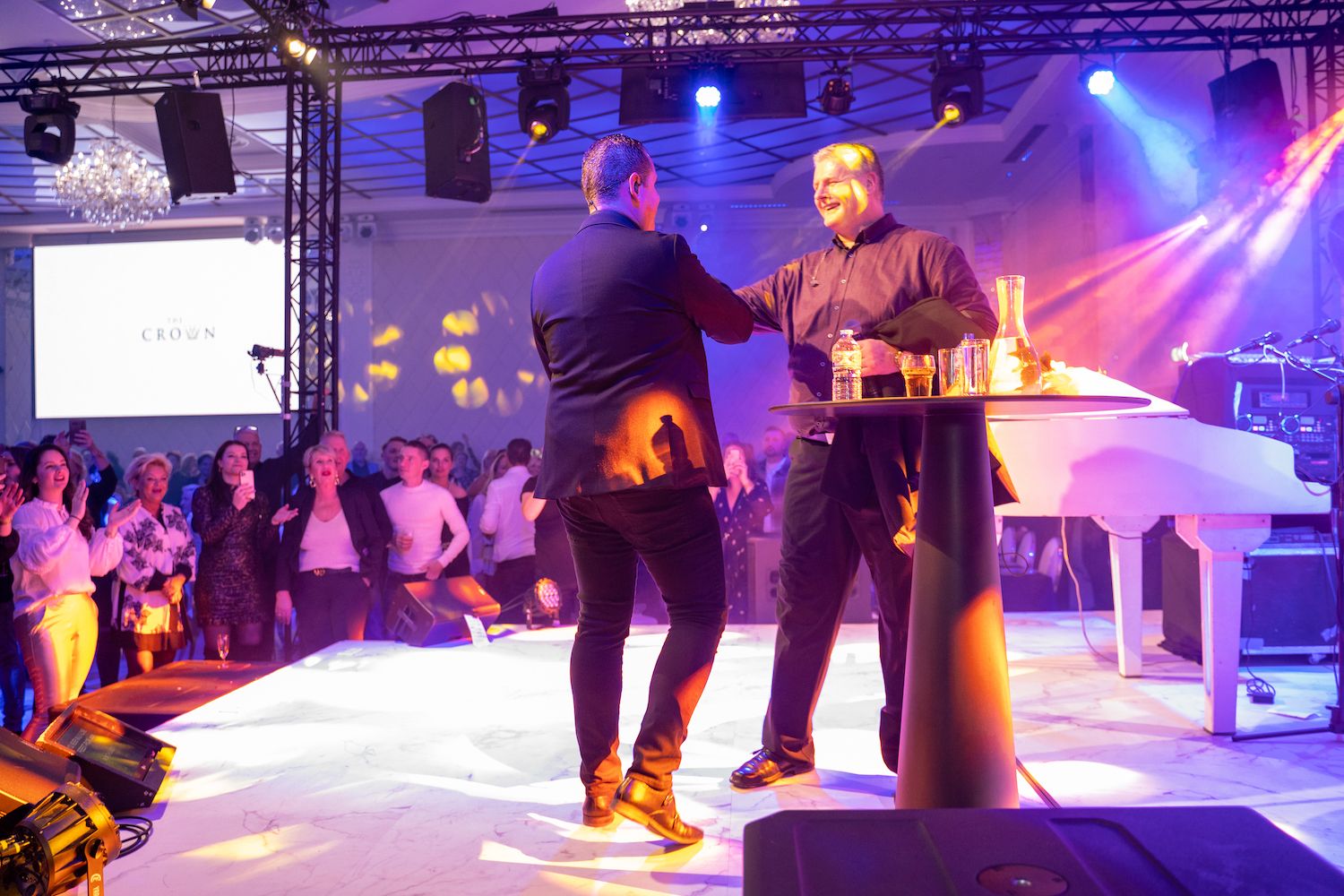 Twee mensen schudden elkaar de hand op een verlicht podium voor een publiek, met een witte vleugel in de buurt.