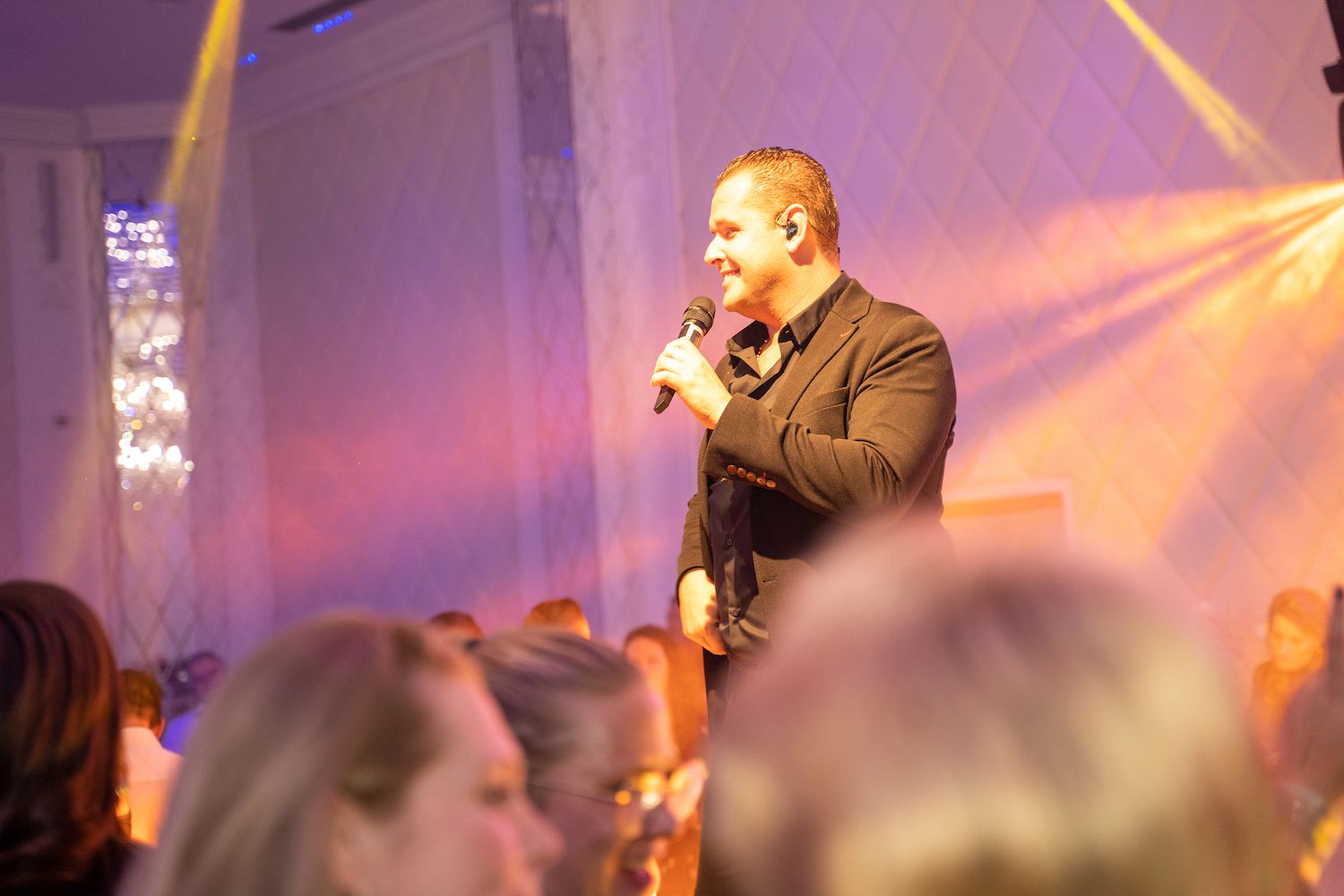 Een man spreekt in een microfoon op een podium met paarse verlichting en een wazig publiek op de voorgrond.