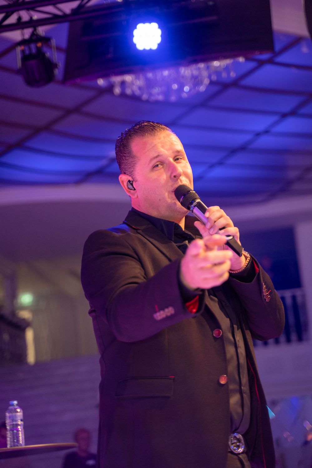 Artiest in een zwart pak zingt in een microfoon op een podium onder paarse verlichting.