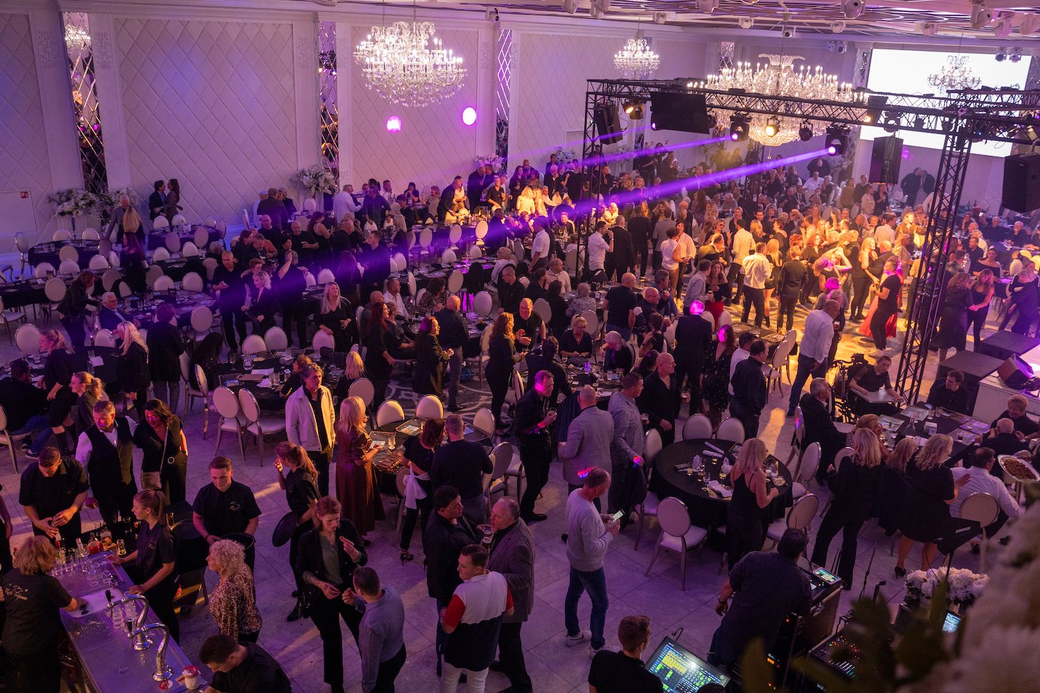 Een drukbezocht binnenfeest met mensen die dansen onder paarse verlichting en kroonluchters.