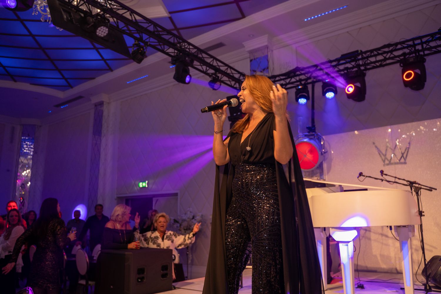 Zangeres treedt op het podium op in een paars verlichte evenementenhal, met een microfoon naast een witte piano.