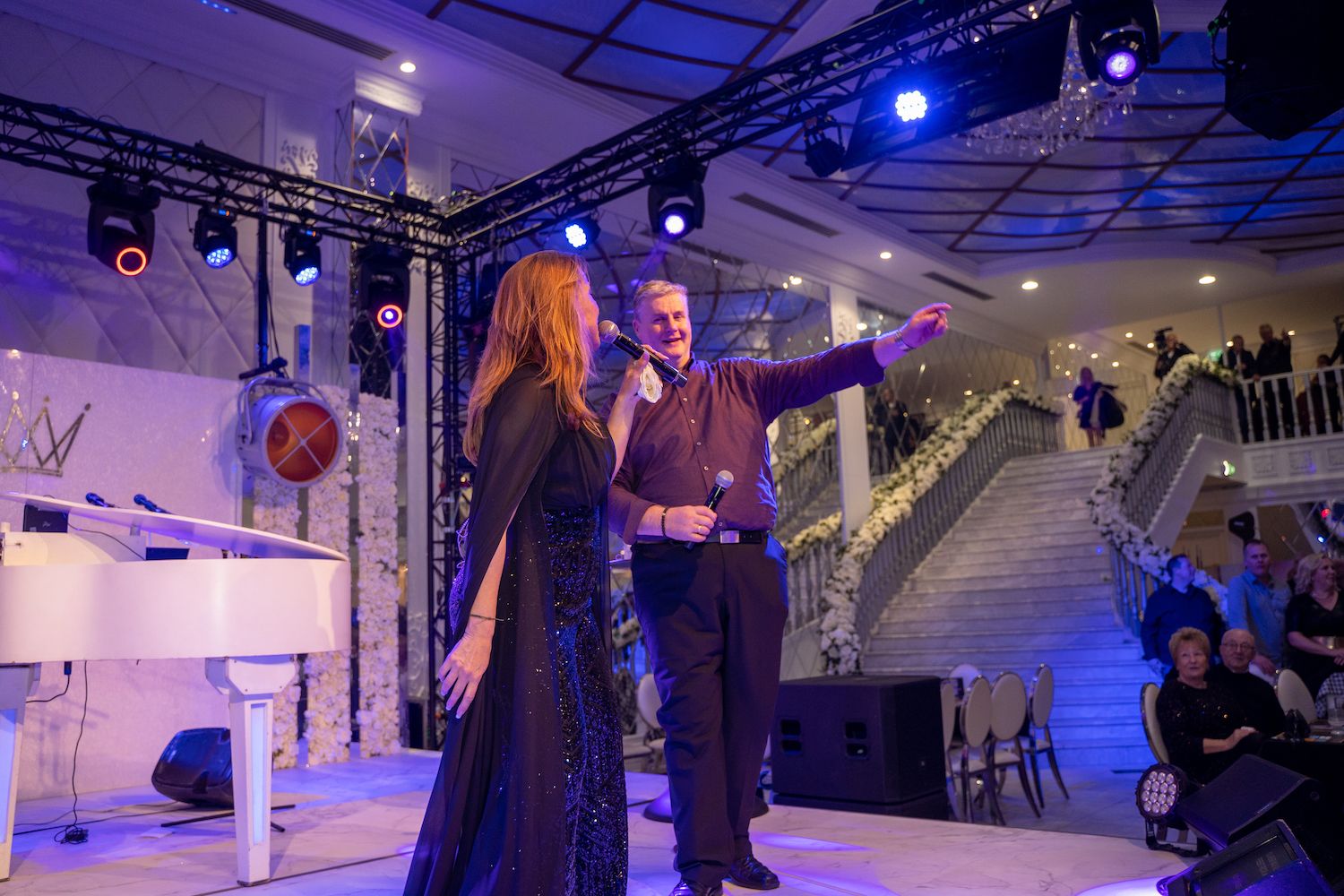 Twee artiesten op een verlicht podium, de een zingt en de ander wijst, in een versierde binnenruimte.