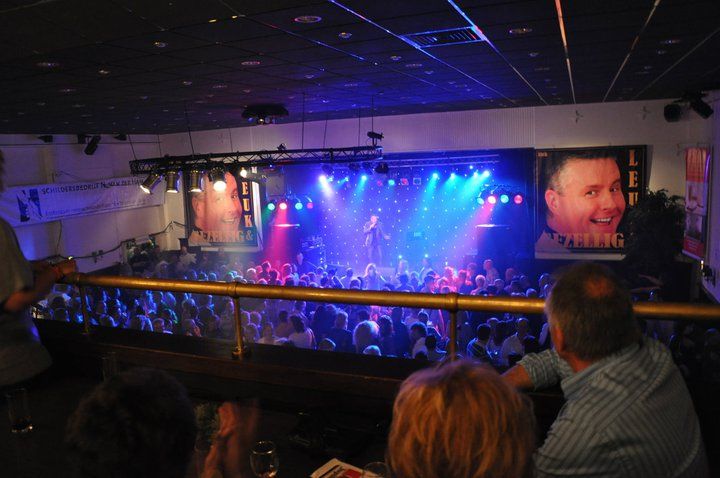 Concertpubliek in een blauwverlichte club, met rechts op een groot scherm een ​​lachend gezicht.