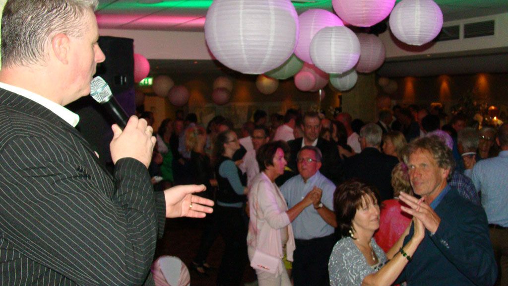 Een man spreekt in een microfoon op een druk feest met hangende lantaarns en gasten die om hem heen zitten en staan.