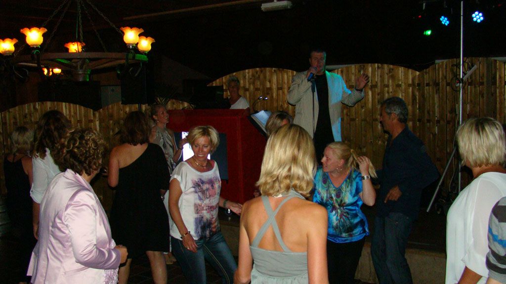 Mensen dansen op een nachtelijk buitenfeest onder hangende lampen, met een muzikant op het podium.