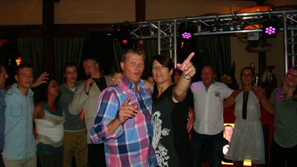 Mensen dansen en poseren op een levendig binnenfeest met kleurrijke lichten, met een man in een geruit overhemd vooraan.