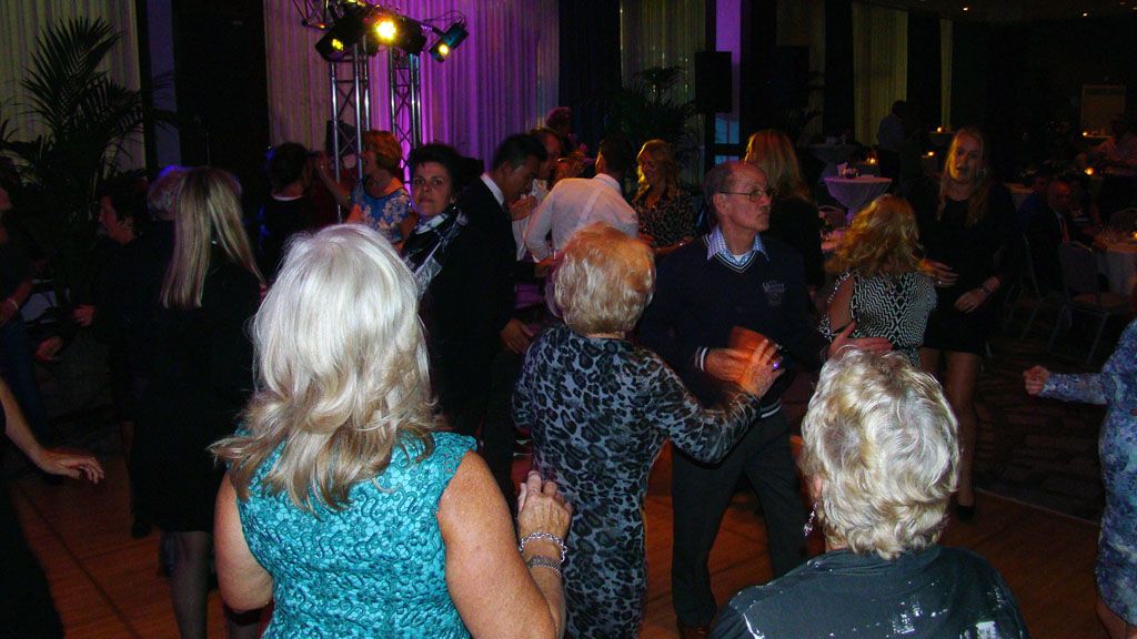 Mensen dansen op een schemerig verlicht feest, met paarse lampen en gasten die op de achtergrond toekijken.