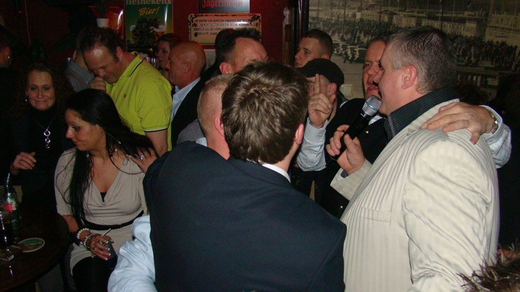 Mensen zingen en dansen op een druk binnenfeest, terwijl een man een microfoon vasthoudt.