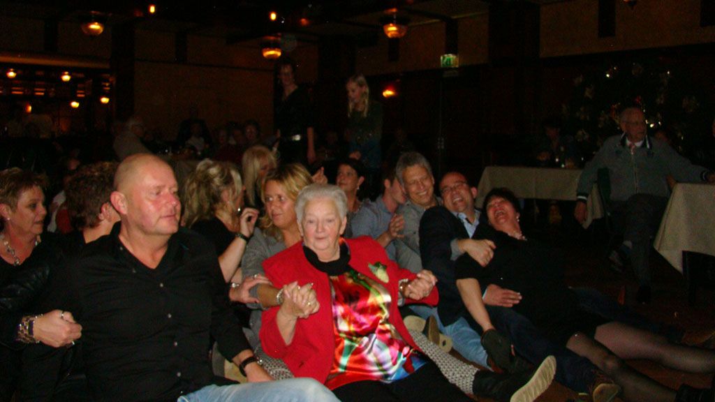Het publiek zit in een schemerig theater, met een oudere vrouw in een rode jas met bloemenprint vooraan.