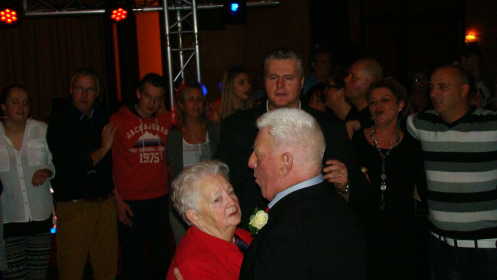 Een ouder stel danst op een schemerig verlicht feest, terwijl gasten op de achtergrond toekijken.