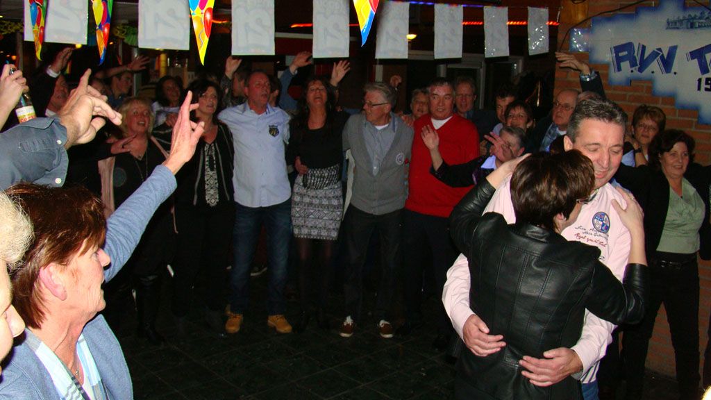 Mensen dansen op een druk binnenfeest met vlaggen en kleurrijke versieringen boven hun hoofden.
