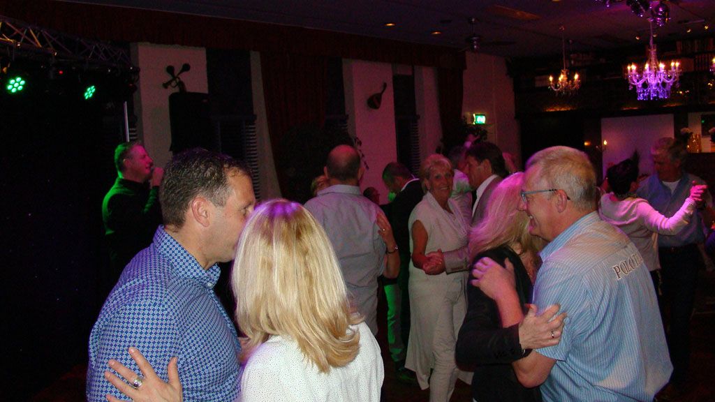 Mensen dansen en praten in een schemerig verlichte feestzaal met paarse en groene lampen.