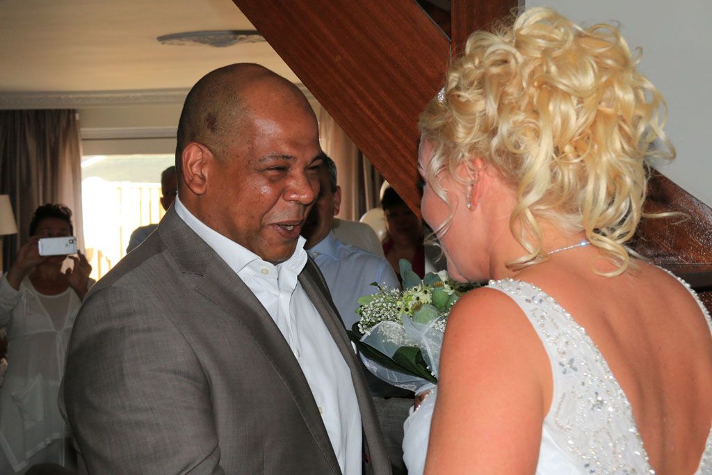 Twee mensen in gesprek op een bruiloftsreceptie, met bloemen en gasten op de achtergrond.