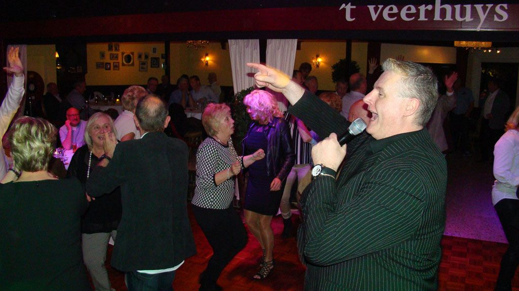 Mensen dansen in een schemerig verlichte club, met op de voorgrond een man in een groen shirt die zijn arm omhoog steekt.