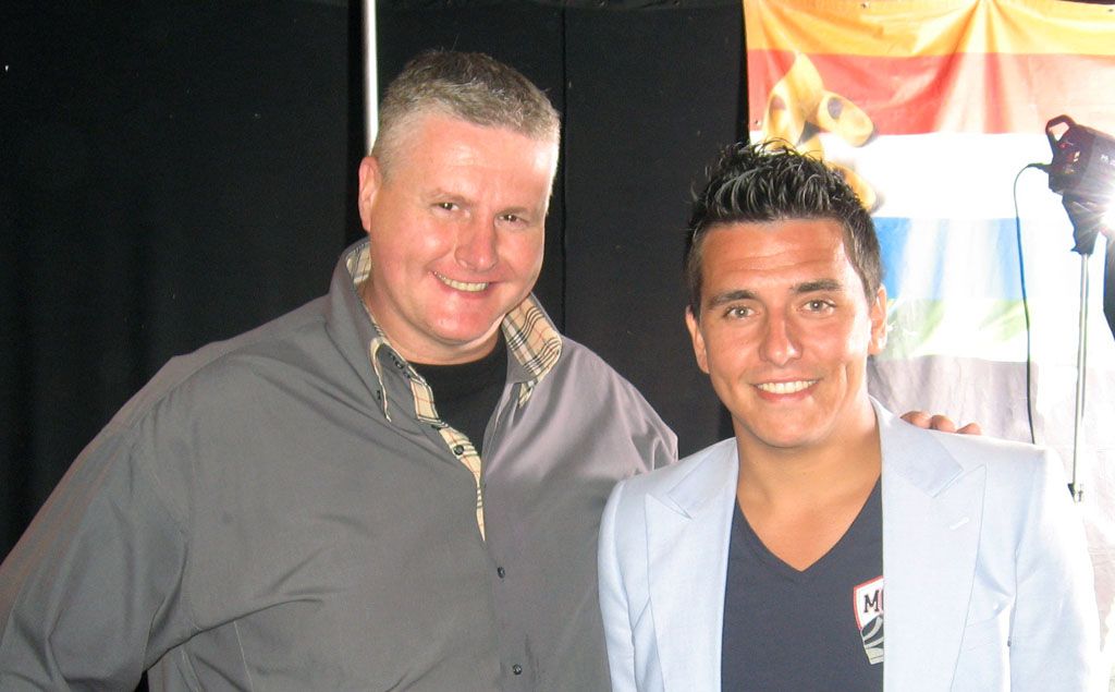 Twee mannen glimlachen tijdens een evenement binnenshuis, de een in een grijs overhemd en de ander in een lichtblauwe blazer.